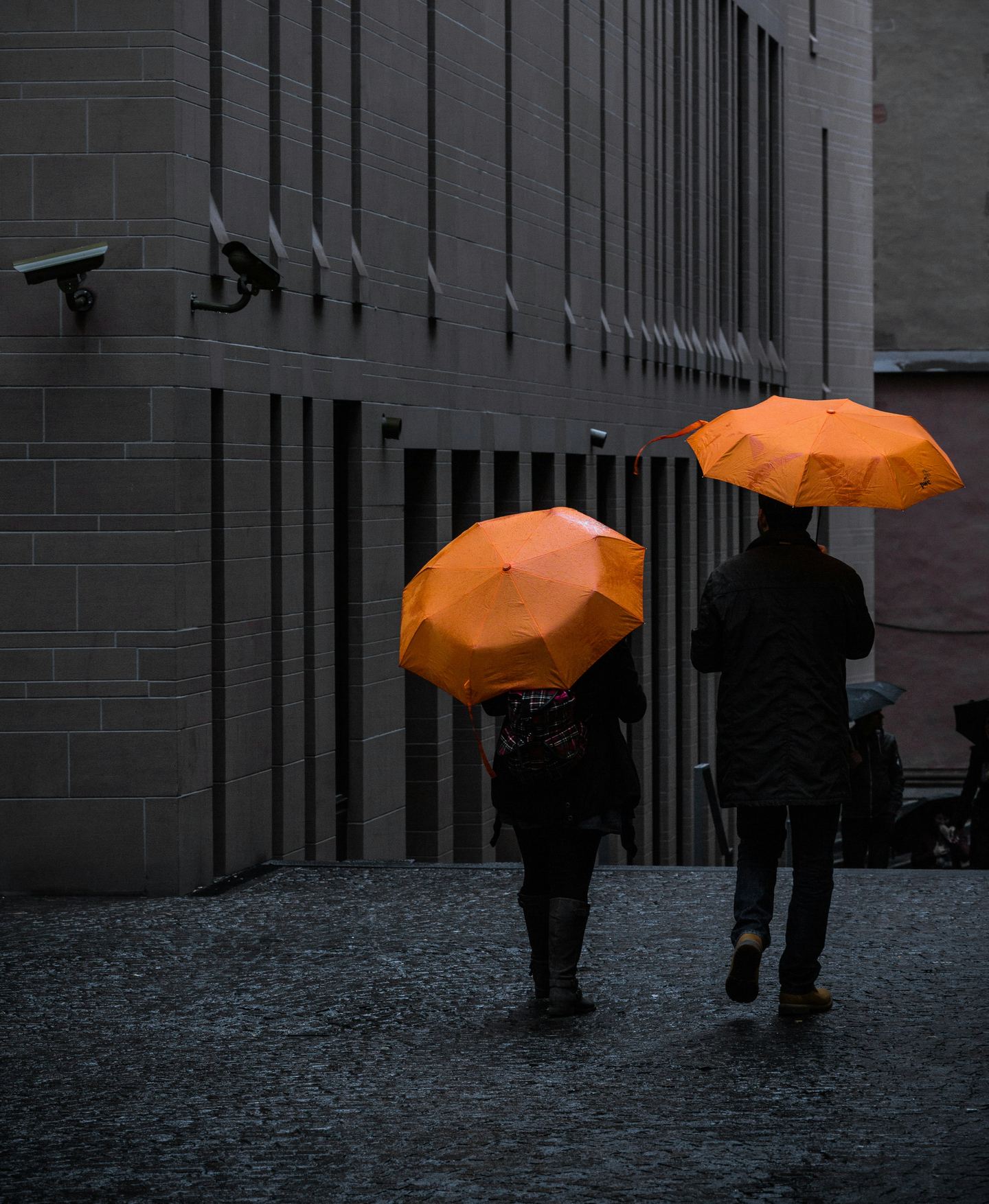 Two people walking with orange umbrellas beside a concrete wall